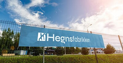 Et blåt banner med hvid tekst, hvor der står "Hegnsfabrikken", hænger på et metalhegn. Himlen er delvist overskyet og solen skinner. Træer og bygninger er synlige i baggrunden, hvilket tyder på et semi-bymiljø, hvor sådanne bannere tilføjer charme til hegn.