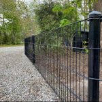 Et sort metalhegn løber langs en grussti omkranset af træer og forskelligt grønt, med Stolpehat 60x40mm GRÅ Plast Pyramide, der dækker hver stolpe. Et lille træskur er delvist synligt i baggrunden, beliggende mellem træerne. Scenen er udendørs og ser ud til at være i landlige omgivelser eller forstæder.