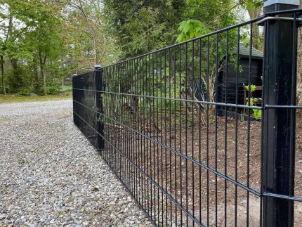Et sort metalhegn løber langs en grussti omkranset af træer og forskelligt grønt, med Stolpehat 60x40mm GRÅ Plast Pyramide, der dækker hver stolpe. Et lille træskur er delvist synligt i baggrunden, beliggende mellem træerne. Scenen er udendørs og ser ud til at være i landlige omgivelser eller forstæder.