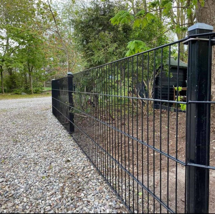 Et sort metalhegn løber langs en grussti omkranset af træer og forskelligt grønt, med Stolpehat 60x40mm GRÅ Plast Pyramide, der dækker hver stolpe. Et lille træskur er delvist synligt i baggrunden, beliggende mellem træerne. Scenen er udendørs og ser ud til at være i landlige omgivelser eller forstæder.