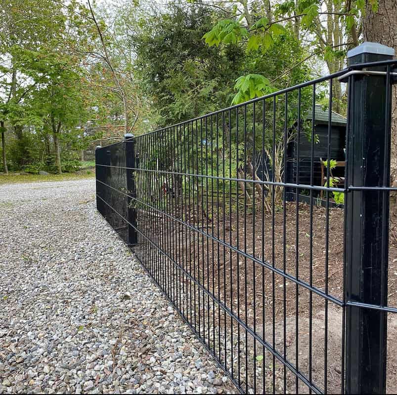 Et sort metalhegn løber langs en grussti omkranset af træer og forskelligt grønt, med Stolpehat 60x40mm GRÅ Plast Pyramide, der dækker hver stolpe. Et lille træskur er delvist synligt i baggrunden, beliggende mellem træerne. Scenen er udendørs og ser ud til at være i landlige omgivelser eller forstæder.