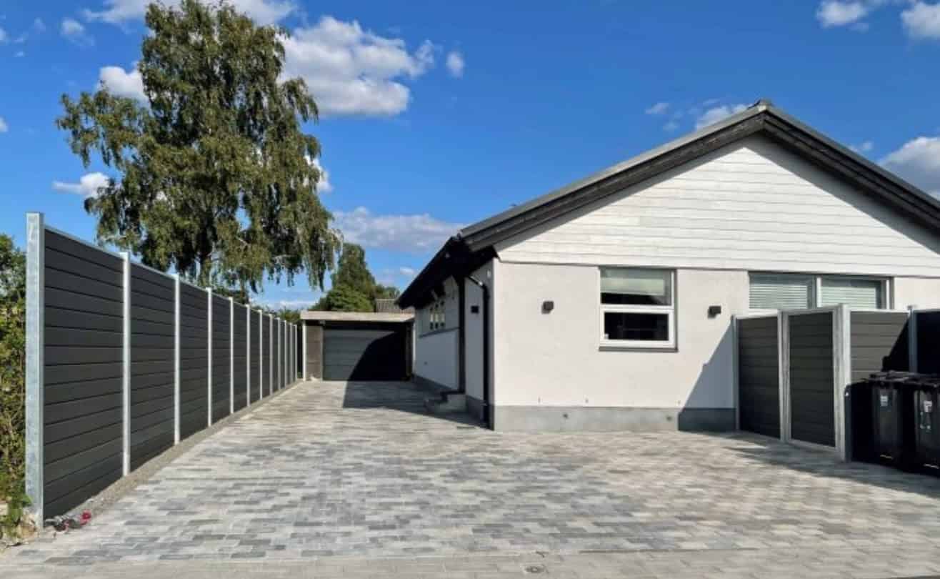 Et moderne etplanshus med hvid facade og mørk tagdækning. Huset ligger for enden af en bred, pænt asfalteret indkørsel, flankeret af høje mørke hegn. I baggrunden er der et stort træ og en klar blå himmel med et par spredte skyer – der viser fordelene ved et hegn udført i WPC / Komposit.