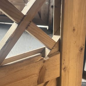 Nærbillede af en Havelåge tæt trykimpr-træstruktur med krydsende diagonale og lodrette bjælker. Sollyset fremhæver de ru kanter, træets årer og splinter, mens baggrunden er sløret.