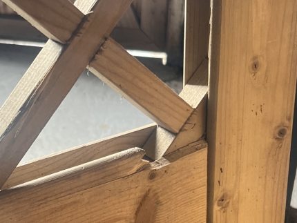 Nærbillede af en Havelåge tæt trykimpr-træstruktur med krydsende diagonale og lodrette bjælker. Sollyset fremhæver de ru kanter, træets årer og splinter, mens baggrunden er sløret.