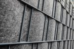Nærbillede af et gråt metalgitter med FLEECELINE Afskærmning rulle 26 m paneler vævet mellem metalstængerne. Gitteret skaber et mønster af rektangulære sektioner, og FLEECELINE-panelerne giver et tekstureret, blødt udseende. Billedet fanger strukturen i en vinkel.