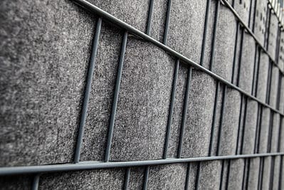 Nærbillede af et gråt metalgitter med FLEECELINE Afskærmning rulle 26 m paneler vævet mellem metalstængerne. Gitteret skaber et mønster af rektangulære sektioner, og FLEECELINE-panelerne giver et tekstureret, blødt udseende. Billedet fanger strukturen i en vinkel.