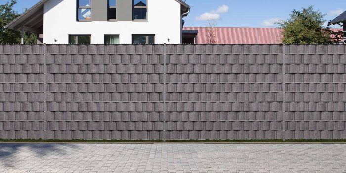 Et højt, gråt privathegn opbygget af metalpaneler og forstærket med FLEECELINE Afskærmning rulle 26 m - Indfletning af Panelhegn står på en murstensbelagt overflade. Bag hegnet ses delvist en hvid bygning med trekantet tag og store vinduer. Træer og en blå himmel kan ses i baggrunden.