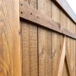 Nærbillede af Havelåge rustikke planker i brun, der viser lodrette planker med synlige naturlige åremønstre. Denne rustikke træhegnslåge har en vandret planke nær toppen og et vinklet støttestykke, der skaber en trekantet form på siden. Det lysebrune træ fremstår forvitret, hvilket tilføjer dets charme.