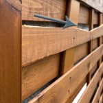 Nærbillede af et plankeværk med en fremtrædende vandret revne, der løber gennem en af dens lameller. En grå pil peger på det beskadigede område og fremhæver den synlige spaltning i træet på denne Havelåge brunmalet. Hegnet har en naturlig finish og fastgøres med søm.