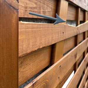 Nærbillede af et plankeværk med en fremtrædende vandret revne, der løber gennem en af dens lameller. En grå pil peger på det beskadigede område og fremhæver den synlige spaltning i træet på denne Havelåge brunmalet. Hegnet har en naturlig finish og fastgøres med søm.