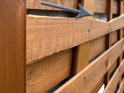 Nærbillede af et plankeværk med en fremtrædende vandret revne, der løber gennem en af dens lameller. En grå pil peger på det beskadigede område og fremhæver den synlige spaltning i træet på denne Havelåge brunmalet. Hegnet har en naturlig finish og fastgøres med søm.