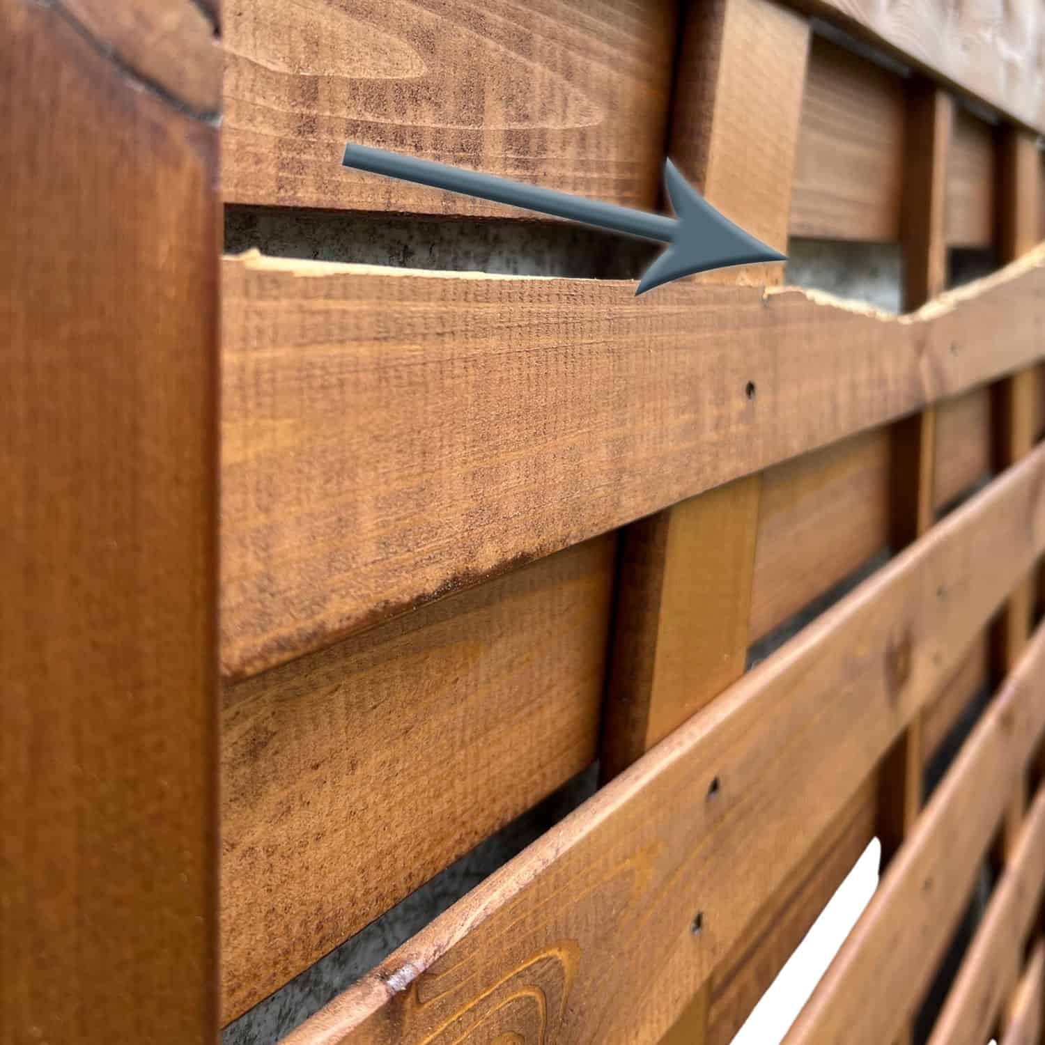 Nærbillede af et plankeværk med en fremtrædende vandret revne, der løber gennem en af dens lameller. En grå pil peger på det beskadigede område og fremhæver den synlige spaltning i træet på denne Havelåge brunmalet. Hegnet har en naturlig finish og fastgøres med søm.