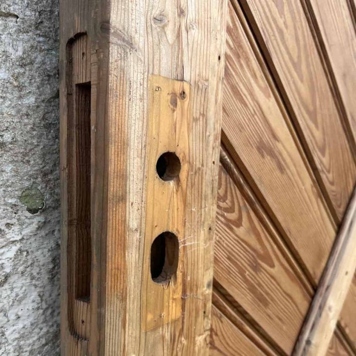 Nærbillede af et Havelåge lærketræ 100x180cm med ufærdige huller, hvor lås og håndtag skal monteres. Det lyse træ fremviser naturlige åremønstre. Porten er delvist åben og afslører en stenmur på venstre side af karmen.