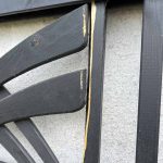 Et nærbillede af Havelåge m/espalier Antracit, et dekorativt træstykke, der minder om et espalier, monteret på en hvid væg. Stykket viser skader med afhugget maling og blotlagt træ langs kanten af rammen. Strukturen har et abstrakt eller geometrisk mønster.