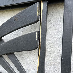 Et nærbillede af Havelåge m/espalier Antracit, et dekorativt træstykke, der minder om et espalier, monteret på en hvid væg. Stykket viser skader med afhugget maling og blotlagt træ langs kanten af rammen. Strukturen har et abstrakt eller geometrisk mønster.