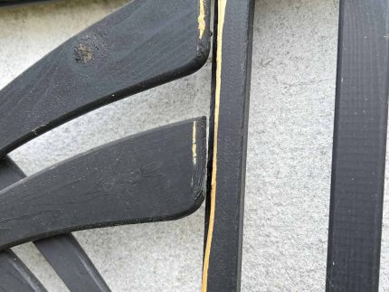 Et nærbillede af Havelåge m/espalier Antracit, et dekorativt træstykke, der minder om et espalier, monteret på en hvid væg. Stykket viser skader med afhugget maling og blotlagt træ langs kanten af rammen. Strukturen har et abstrakt eller geometrisk mønster.