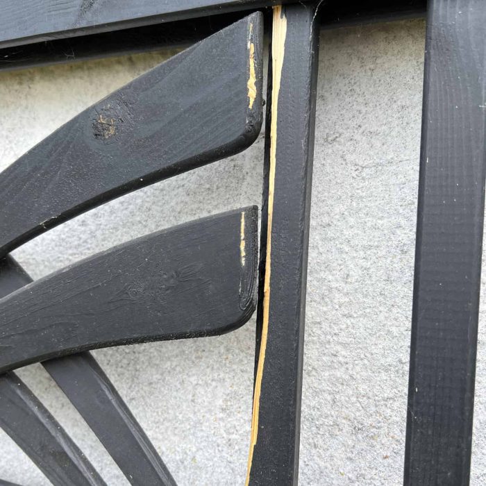 Et nærbillede af Havelåge m/espalier Antracit, et dekorativt træstykke, der minder om et espalier, monteret på en hvid væg. Stykket viser skader med afhugget maling og blotlagt træ langs kanten af rammen. Strukturen har et abstrakt eller geometrisk mønster.