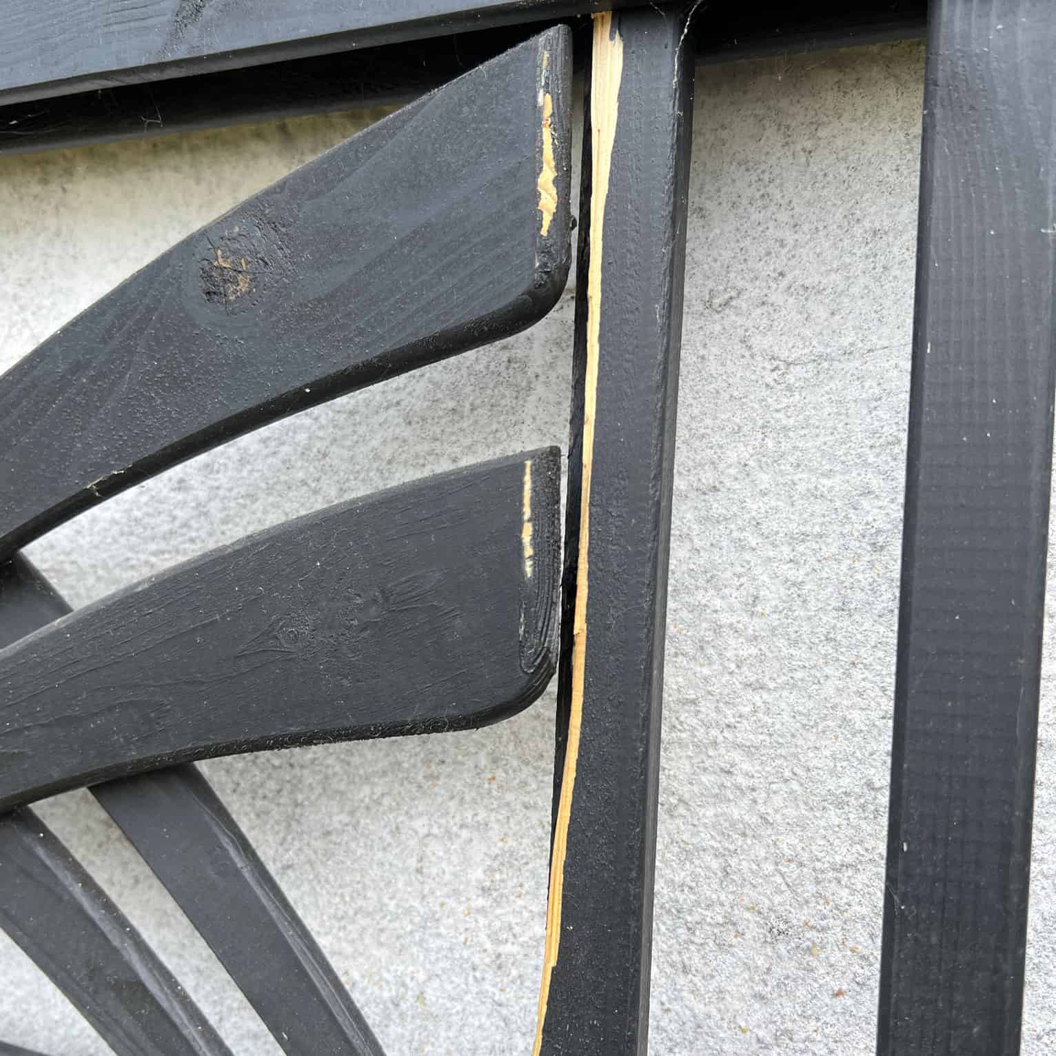 Et nærbillede af Havelåge m/espalier Antracit, et dekorativt træstykke, der minder om et espalier, monteret på en hvid væg. Stykket viser skader med afhugget maling og blotlagt træ langs kanten af rammen. Strukturen har et abstrakt eller geometrisk mønster.