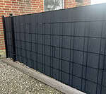 Et moderne, sort panelhegn med et gitterdesign står ved siden af en murstensbygning. Jorden er dækket af grus, og FLEECELINE Afskærmning rulle 26 m - Indfletning af Panelhegn væver sig gennem de lodrette og vandrette stænger og forstærker de firkantede mønstre. En træplanke beklæder bunden af hegnet.