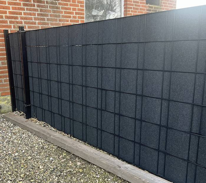 Et moderne, sort panelhegn med et gitterdesign står ved siden af en murstensbygning. Jorden er dækket af grus, og FLEECELINE Afskærmning rulle 26 m - Indfletning af Panelhegn væver sig gennem de lodrette og vandrette stænger og forstærker de firkantede mønstre. En træplanke beklæder bunden af hegnet.