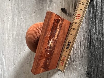 En Stolpehat m/kugle brun træ er placeret ved siden af en lineal, der viser, at den er cirka 10 cm lang, alt sammen sat på en vejrbidt, malet træoverflade.