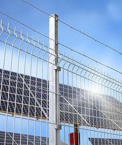 Et højt metalhegn med pigtråd på toppen står mod en klar blå himmel. Solpaneler i baggrunden antyder et solenergianlæg. Sollys skinner af Panelhegn 3D 5/5mm - HF PREMIUM Varmgalvaniseret HDG og paneler.