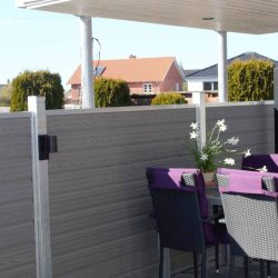 En udendørs terrasse med overdækket tag har en spiseplads med et bord omgivet af lilla og sorte stole. På bordet stilles en potteplante med hvide blomster. Terrassen er omsluttet af gråt træpanel hegn udført i slankt Kompositbræt 25mm - WPC Træstruktur, der giver en naturskøn udsigt over nabohuse og grønne områder.