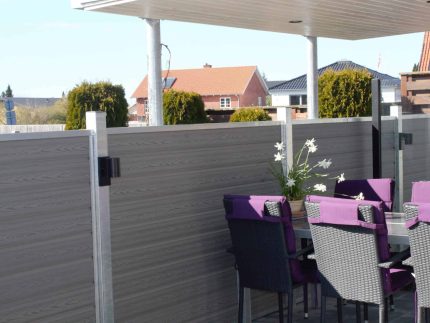 En udendørs terrasse med overdækket tag har en spiseplads med et bord omgivet af lilla og sorte stole. På bordet stilles en potteplante med hvide blomster. Terrassen er omsluttet af gråt træpanel hegn udført i slankt Kompositbræt 25mm - WPC Træstruktur, der giver en naturskøn udsigt over nabohuse og grønne områder.