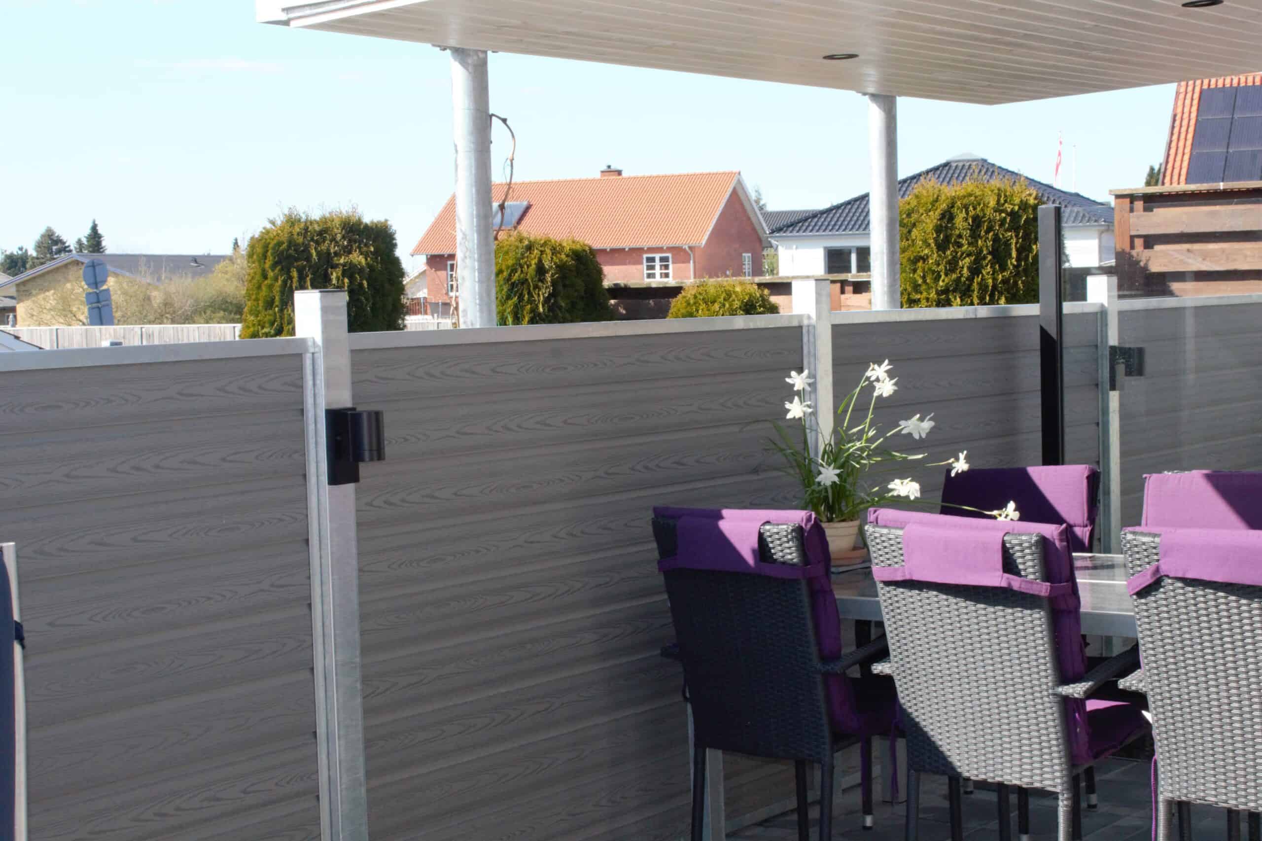 En udendørs terrasse med overdækket tag har en spiseplads med et bord omgivet af lilla og sorte stole. På bordet stilles en potteplante med hvide blomster. Terrassen er omsluttet af gråt træpanel hegn udført i slankt Kompositbræt 25mm - WPC Træstruktur, der giver en naturskøn udsigt over nabohuse og grønne områder.