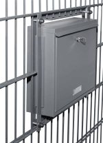 En Postkasse til indbygning i panelhegn - Antracit 7016 Måtte er monteret på et metalhegn. Den grå, aflåselige postkasse har en lille spalte til aflevering af post og et nøglehul til sikring af indholdet.
