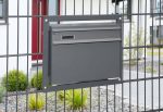 En moderne Postkasse til indbygning i panelhegn - Antracit 7016 Mat er problemfrit indbygget i et lækkert metalhegn foran et moderne hus med rød dør. Jorden er dækket af grus, små planter og buske, hvilket skaber et pænt og minimalistisk uderum.