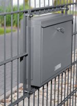 En Postkasse til indbygning i panelhegn - Antracit 7016 Mat er monteret på en metalpanelhegn. Postkassen har en lås på fronten og en hul til post øverst. Rundt om hegnet er en asfalteret sti og et pænt trimmet græsareal med nogle sten nær bunden af hegnet.