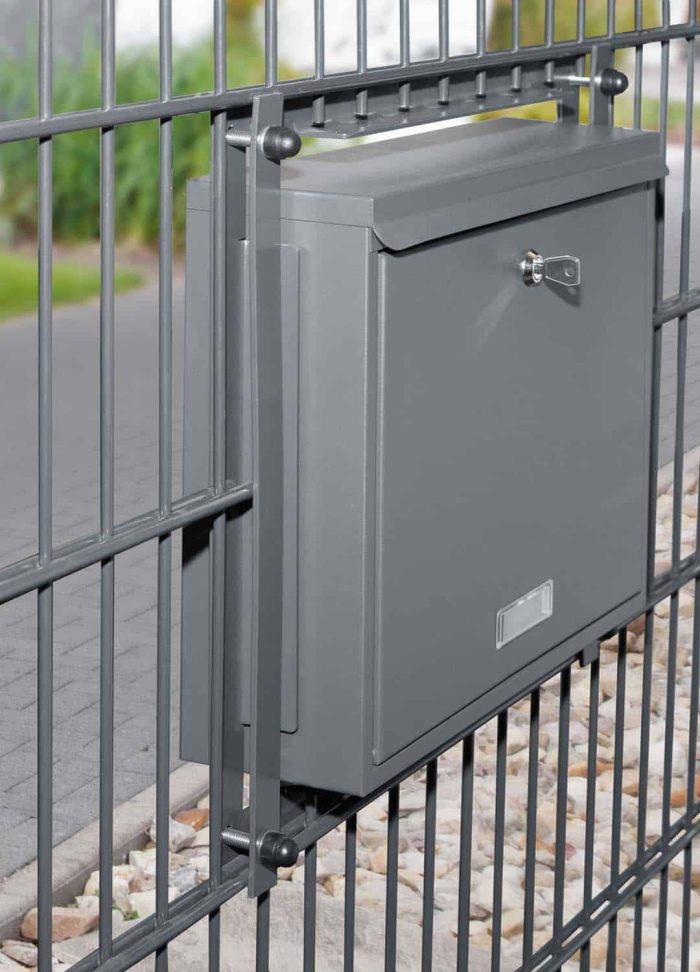 En Postkasse til indbygning i panelhegn - Antracit 7016 Mat er monteret på en metalpanelhegn. Postkassen har en lås på fronten og en hul til post øverst. Rundt om hegnet er en asfalteret sti og et pænt trimmet græsareal med nogle sten nær bunden af hegnet.