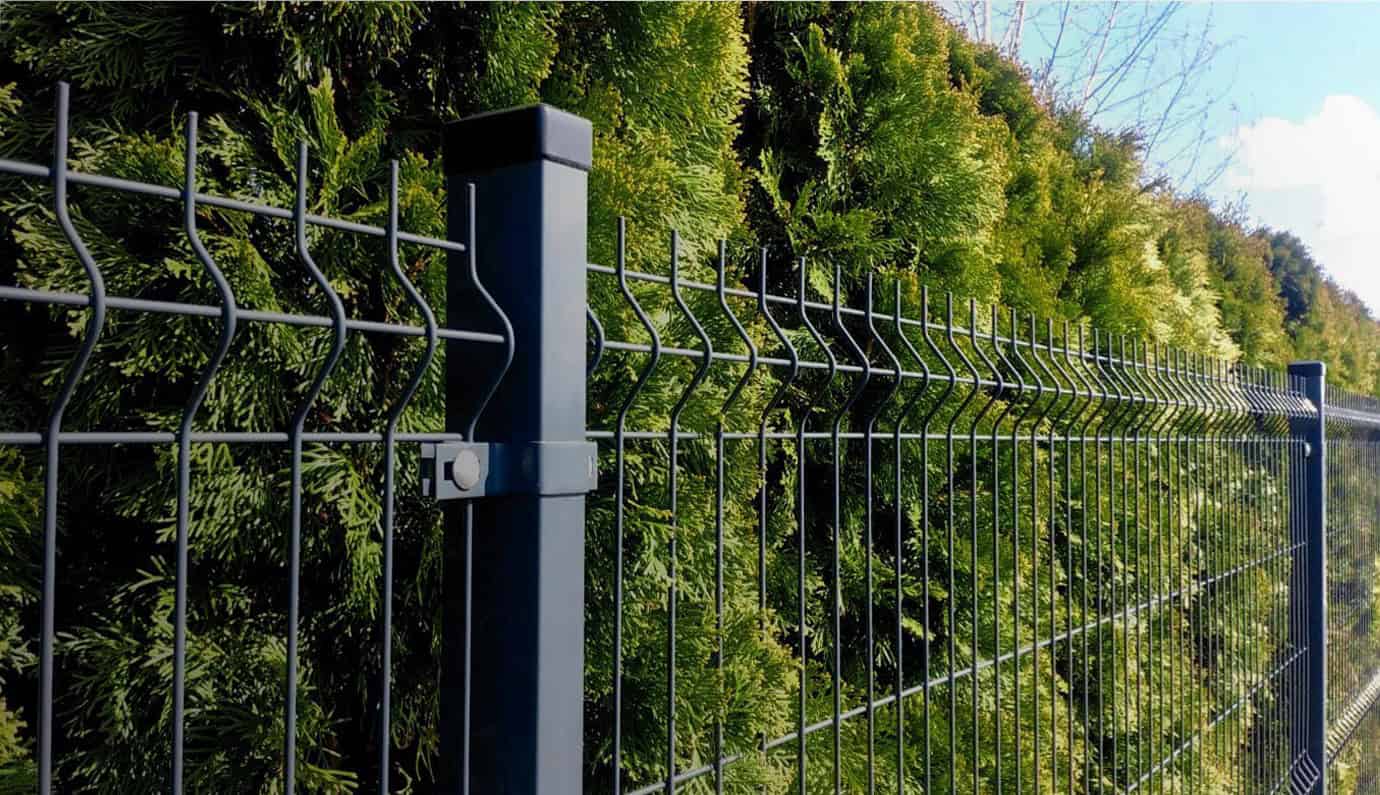 Et sort metalhegn fra Hegnsfabrikken løber parallelt med en tæt, grøn hæk. Hegnet har lodrette stænger forbundet med vandrette zigzag-tråde. Bag hækken ses en klar blå himmel med nogle skyer. Scenen, der viser Slider 5's håndværk, er godt oplyst af dagslys.