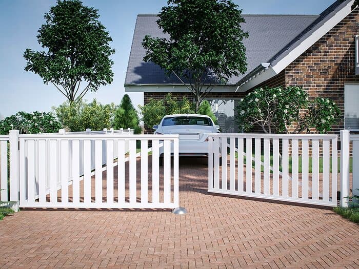 En hvid bil er parkeret i en indkørsel, der fører til et hus med muret udvendigt. Indkørslen på 400x90cm er brolagt med røde mursten og flankeret af "Rækværket" Dobbeltlåge i Plast dobbeltporte, 400x90cm (BRED), hvorfra der løber et hvidt stakit. Haven har grønne buske og høje træer, sat mod en klar blå himmel.