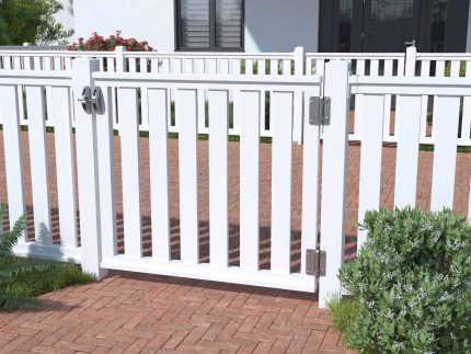 En hvid stakit-låge står åben på en gangbro af røde mursten. Den venstre side af porten er sikret med en lås, mens den højre side har metalhængsler. Omkring området er frodige grønne buske og planter. Husets hvide vægge og vinduer danner en charmerende kulisse til dette 400x90cm Dobbeltlåge i Plast "Rækværk" (BRED), der minder om klassisk design.