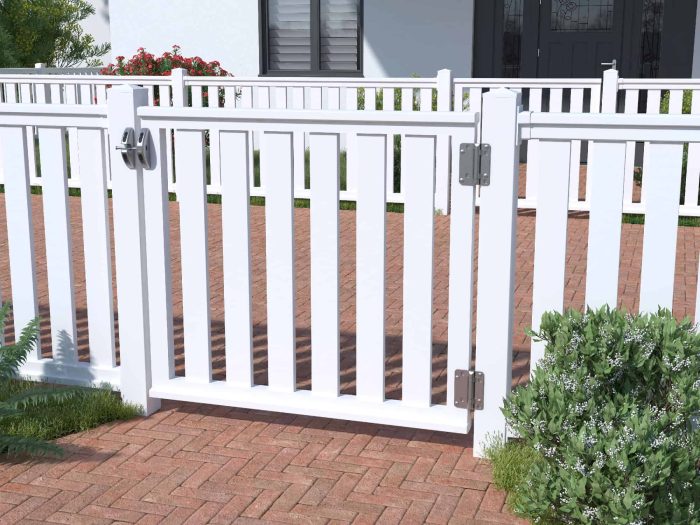 En hvid stakit-låge står åben på en gangbro af røde mursten. Den venstre side af porten er sikret med en lås, mens den højre side har metalhængsler. Omkring området er frodige grønne buske og planter. Husets hvide vægge og vinduer danner en charmerende kulisse til dette 400x90cm Dobbeltlåge i Plast "Rækværk" (BRED), der minder om klassisk design.