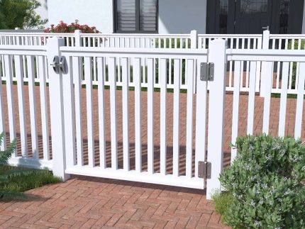 En hvid hegnslåge, nærmere bestemt Hegnslåge i Plast "gelænder" 100x90cm (SMAL), står åben på en rødbrun murstensgang. Hegnet omkranser et pænt anlagt område med grønne buske og planter. Huset i baggrunden har en mørk hoveddør og et vindue med lukkede skodder.