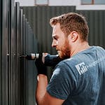 En mand i en T-shirt med "Hege" skrevet på bagsiden bruger en boremaskine til at samle et metalpanelhegn i udendørs omgivelser. Han virker fokuseret på sit arbejde, dedikeret til at bygge robuste hegn og låger til området.