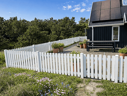 Et moderne hus med solpaneler, et sort ydre og en gårdhave omkranset af Stakithegn i Plast "PROFEXline" 180x80cm (LIGE). Potteplanter pynter på terrassen, og der er træer og blomster i haven under en blå himmel.