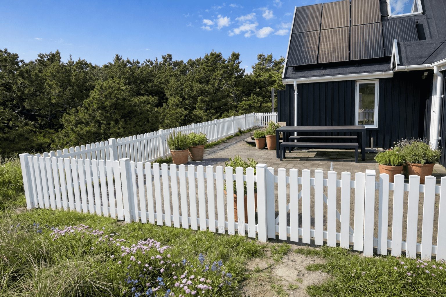 Et moderne hus med solpaneler, et sort ydre og en gårdhave omkranset af Stakithegn i Plast "PROFEXline" 180x80cm (LIGE). Potteplanter pynter på terrassen, og der er træer og blomster i haven under en blå himmel.