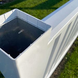 Nærbillede af en udendørs, hvid, rektangulær plaststolpe, identificeret som Stolpe i Plast 120x120mm med alukerne HVID, med den ene ende åben, hvilket afslører det hule indre. Stolpen er fastgjort til et hvidt panel, der ser ud til at være en del af et hegn. Jorden har græs og snavs, og sollys kaster skygger på genstandene.