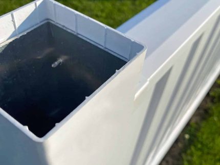 Nærbillede af en udendørs, hvid, rektangulær plaststolpe, identificeret som Stolpe i Plast 120x120mm med alukerne HVID, med den ene ende åben, hvilket afslører det hule indre. Stolpen er fastgjort til et hvidt panel, der ser ud til at være en del af et hegn. Jorden har græs og snavs, og sollys kaster skygger på genstandene.