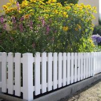 En Stakithegn i Plast "PROFEXline" 180x80cm (LIGE) omgiver et havebed med høje gule og lilla blomster, sat op mod grønne områder og en lys bygning.