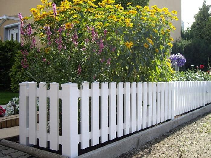 En Stakithegn i Plast "PROFEXline" 180x80cm (LIGE) omgiver et havebed med høje gule og lilla blomster, sat op mod grønne områder og en lys bygning.