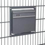 En slank, antracit 7016 mat Postkasse til indbygning i panelhegn er monteret i et metalhegn. Postkassen har en smal spalte til post og en hængslet klap nedenunder. Hegnet har et gittermønster, og kassen fastgøres med fire skruer, en i hvert hjørne af monteringsrammen.