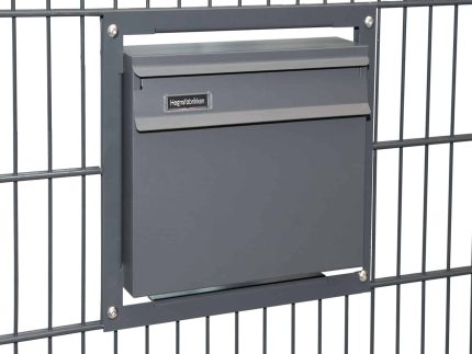 En slank, antracit 7016 mat Postkasse til indbygning i panelhegn er monteret i et metalhegn. Postkassen har en smal spalte til post og en hængslet klap nedenunder. Hegnet har et gittermønster, og kassen fastgøres med fire skruer, en i hvert hjørne af monteringsrammen.