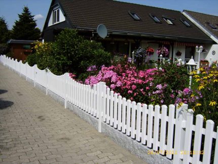 Et malerisk hus med mørkt tag og hvide lister er omkranset af et malerisk Stakithegn i Plast "PROFEXline" 180x80/70cm (BUE NED) hvidt stakithegn. En række farverige blomster, herunder lyserøde, lilla og gule blomster, langs gangbroen ved siden af hegnet. Forgrunden har en brostensbelagt sti.