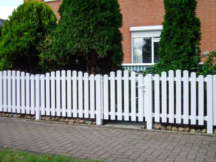 Et hvidt stakit, modelleret i Stakithegn i Plast "PROFEXline" 180x90/80cm (BUE OP) stil, adskiller et fortov fra et rødt murstenshus med et hvidt indrammet vindue. Høje, grønne buske langs det indre af PROFESSline-hegnet, som har en lille låge og en række sten i bunden.