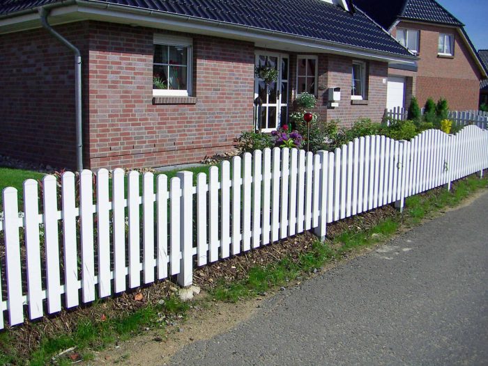 Et malerisk murstenshus med mørkt tegltag ses i baggrunden. Forhaven har en frodig have med forskellige planter og blomster. Et rent Stakithegn i Plast "PROFEXline" 180x90/80cm (BUE OP) hvidt stakit kanter ejendommen, der adskiller den fra en brolagt gade. Himlen er klar og blå.